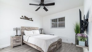 Une chambre à coucher avec un cadre de lit en bois, un ventilateur de plafond, une table de chevet avec une lampe, un téléviseur fixé au mur et une fenêtre avec des stores.