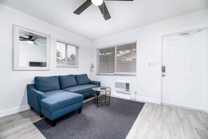 A modern living room with a blue sofa, a small round table, a ceiling fan, and a white door. - Comfy Condos @ Middle River - #3 (Fort Lauderdale)