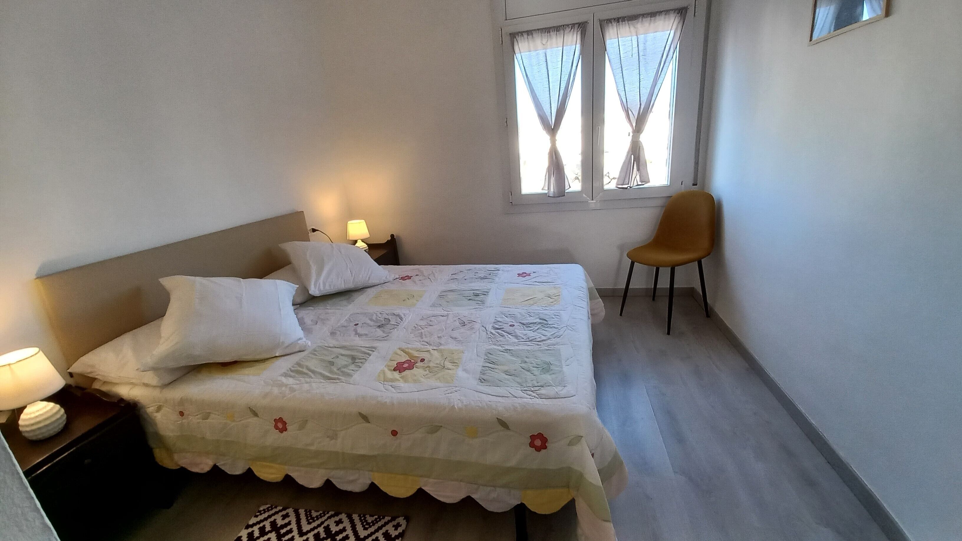 A bedroom with a bed, two pillows, a chair, a nightstand with a lamp, and a window with curtains.