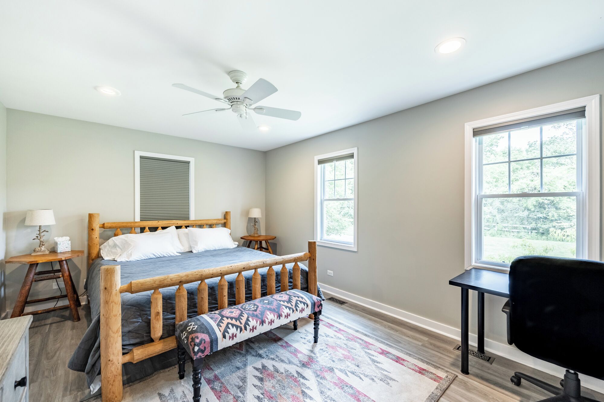 A bedroom with a bed, a wooden bench, a desk, and two windows.