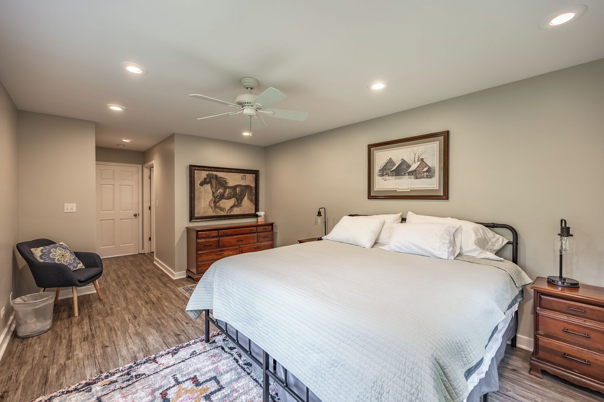 A bedroom with a bed, a dresser, a chair, a ceiling fan, and two framed pictures on the wall.