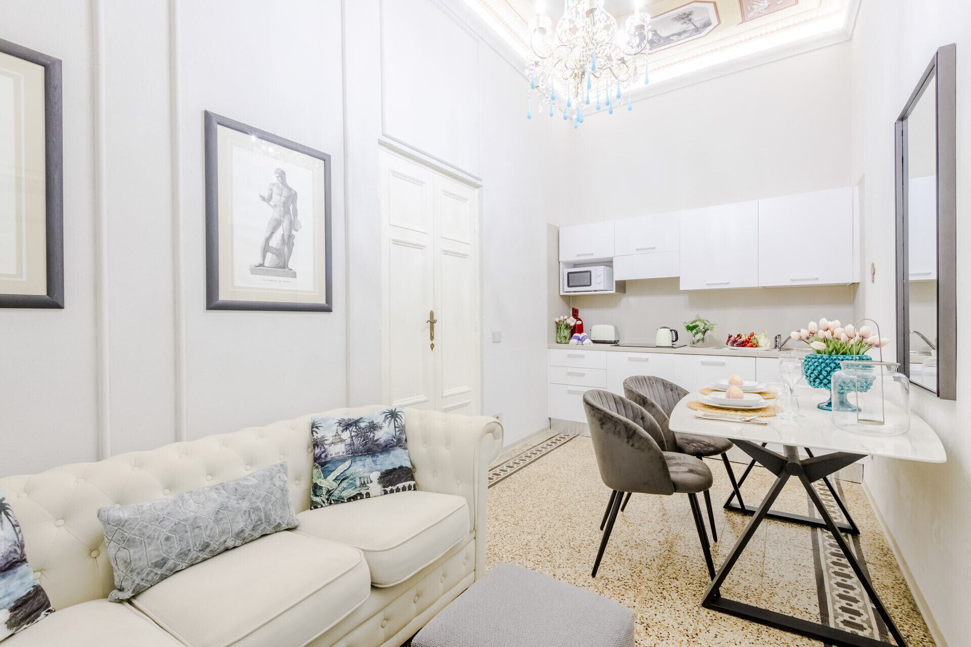 Ein modernes Wohnzimmer mit einem weißen Sofa, einem Esstisch mit Stühlen, einer Küchenzeile mit weißen Schränken und einer Kristallleuchter.