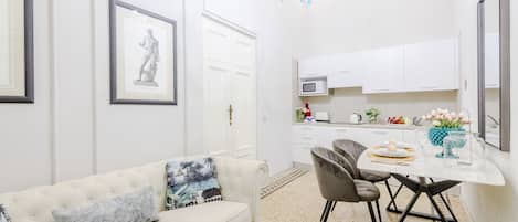 A modern living room with a white sofa, a dining table with chairs, a kitchen area with white cabinets, and a chandelier.