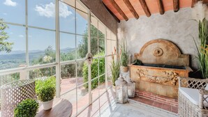 A room with large windows offering a view of the countryside. There is a rustic stone fireplace with a decorative headpiece, surrounded by potted plants and a wicker chair. - Villa Lina, Luxury Farmhouse with Pool and Amazing View close to Lucca Town Centre (San Concordio di Moriano)