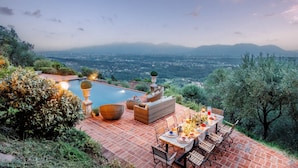 A patio with a pool, outdoor furniture, and a table set for dining, overlooking a scenic landscape with mountains in the distance. - Villa Lina, Luxury Farmhouse with Pool and Amazing View close to Lucca Town Centre (San Concordio di Moriano)