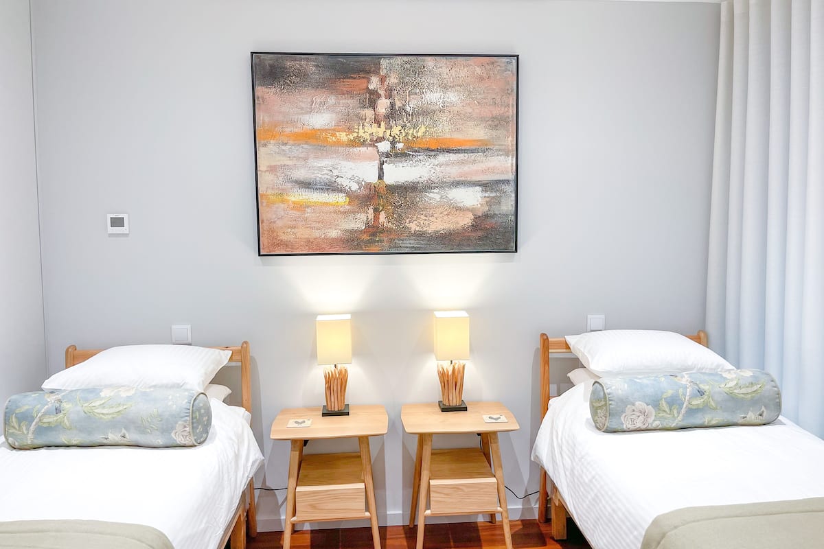 A hotel room with two single beds, each with a floral pillow, a bedside table with a lamp, and a large abstract painting on the wall.