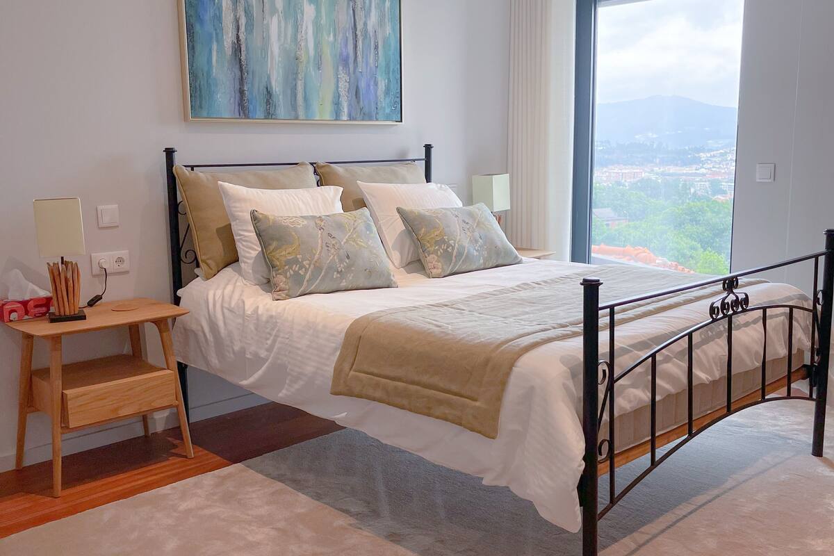 A modern bedroom with a large bed, a bedside table, a nightstand, a painting, and a view of a cityscape through a large window.