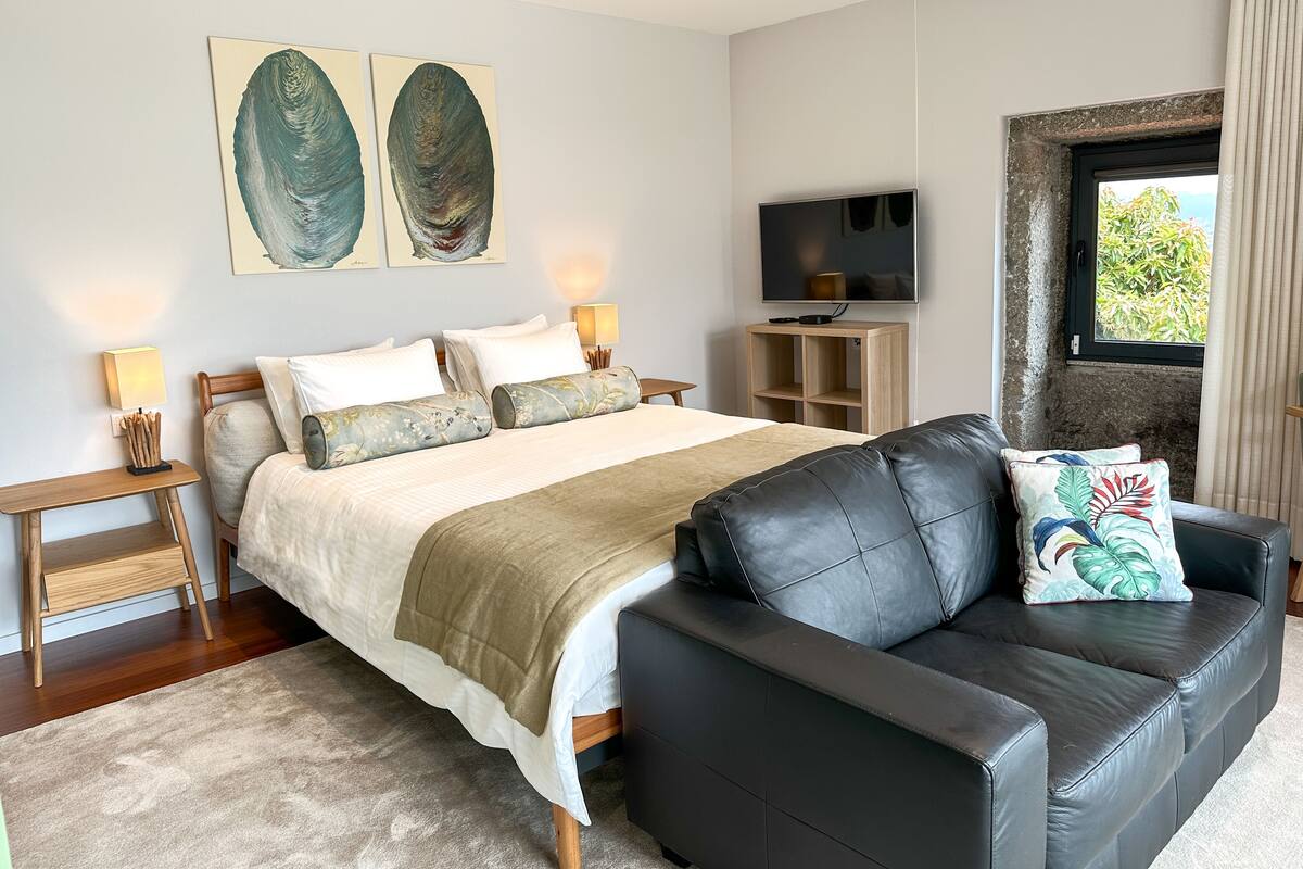 A modern bedroom with a large bed, a black leather sofa, a TV, and artwork on the wall.