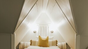 A bedroom with a sloped ceiling, a bed with white bedding and a yellow pillow, and a small window with curtains. - NEW! Off to the races: Stay at the spacious Woodlawn Tudor, close to Oaklawn! (Hot Springs)