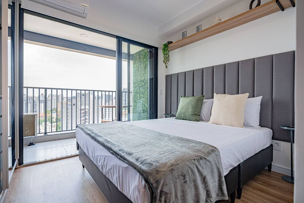 Une chambre moderne avec un grand lit, un balcon offrant une vue sur la ville et une étagère en bois au-dessus du lit.