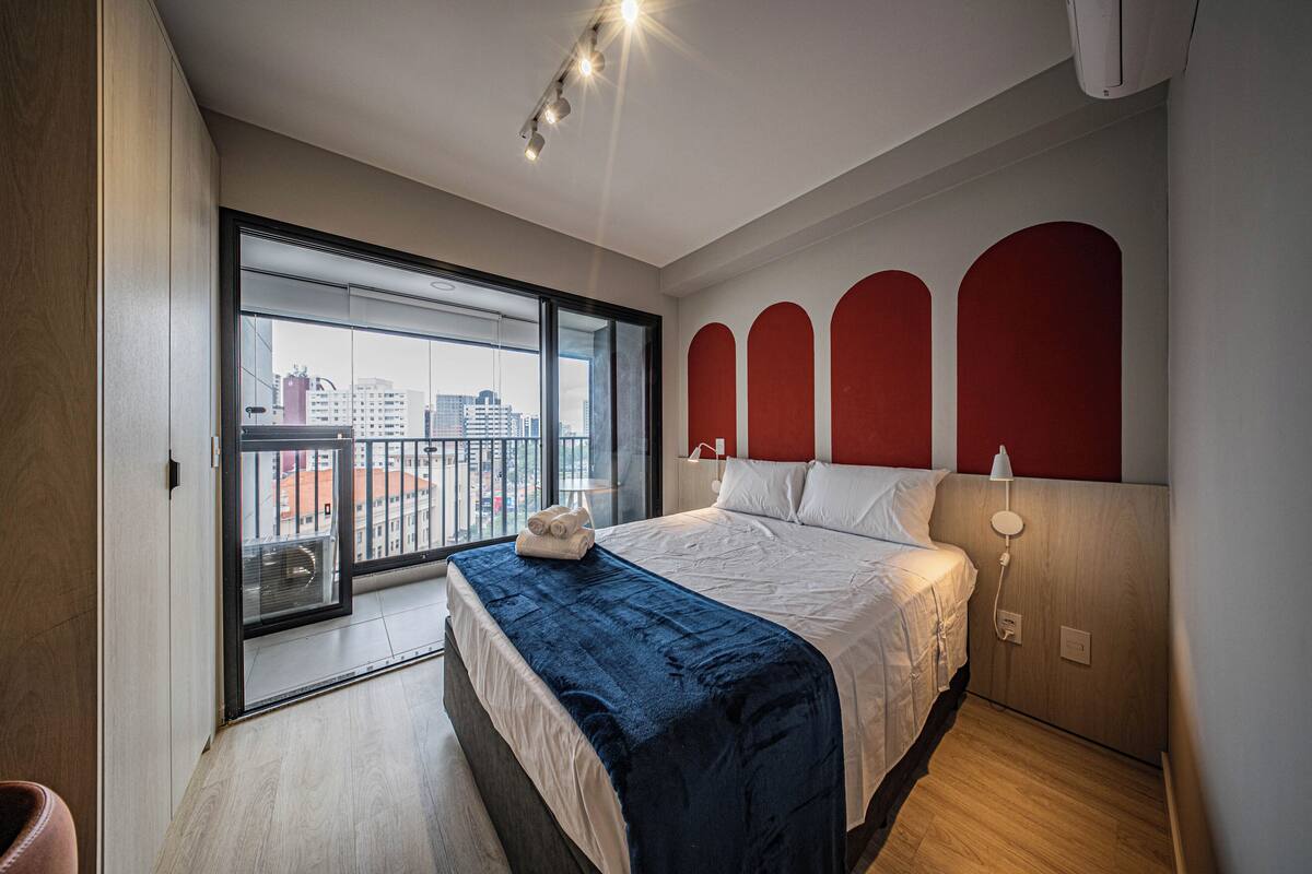Une chambre d’hôtel moderne avec un grand lit, un balcon offrant une vue sur la ville et un mur d’accent rouge.