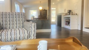 Un salon avec un fauteuil à motifs, une table basse sur laquelle se trouvent une tasse et un livre, et une vue sur la cuisine.
