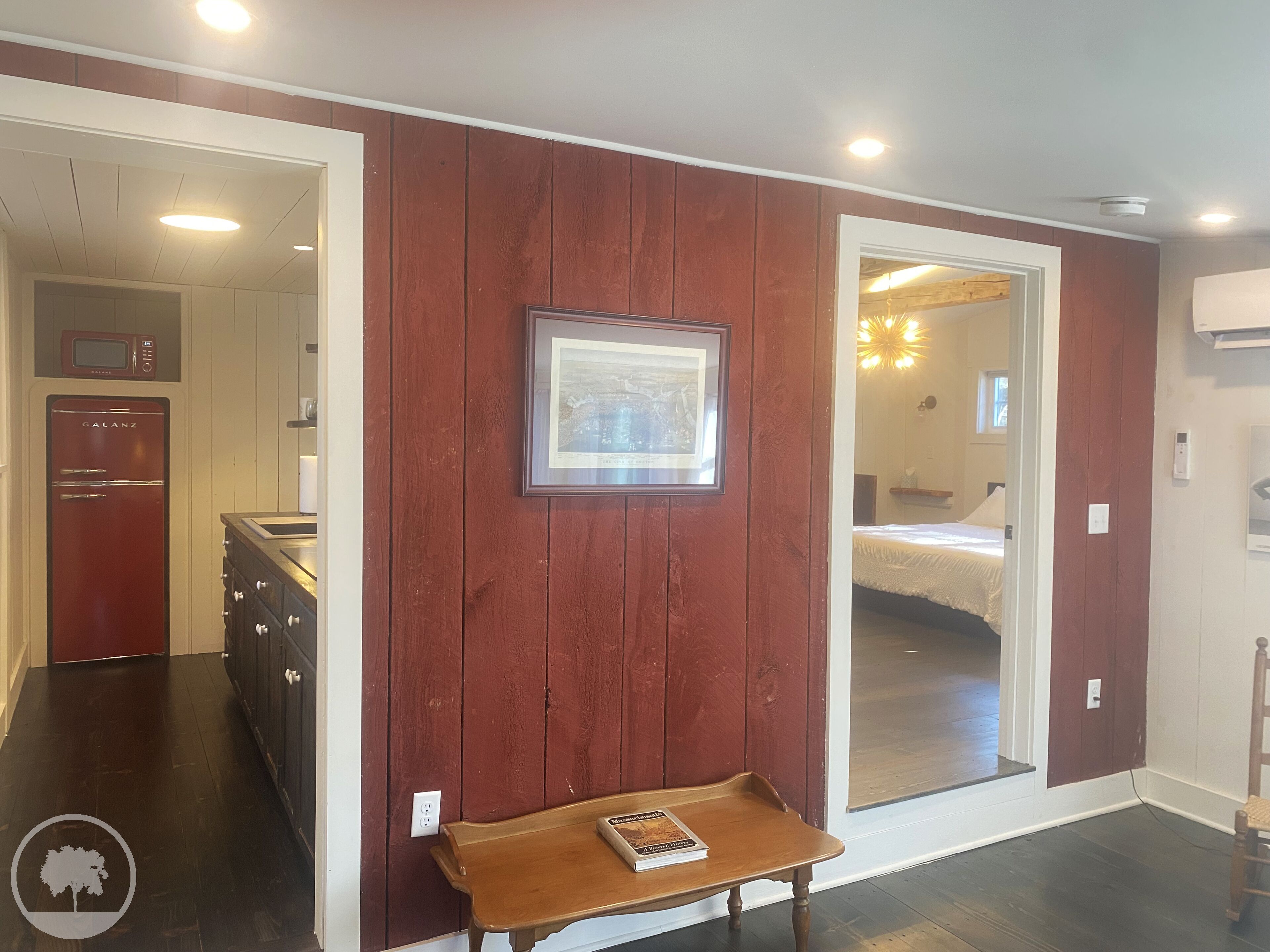 A room with a wooden floor, a small table with a book, a framed picture on the wall, and a door leading to another room.