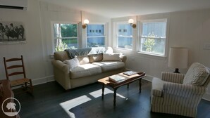 Un salon avec un canapé beige, un fauteuil à rayures, une table basse en bois et une lampe. Il y a deux fenêtres avec des rideaux, une lampe fixée au mur et une affiche accrochée au mur.