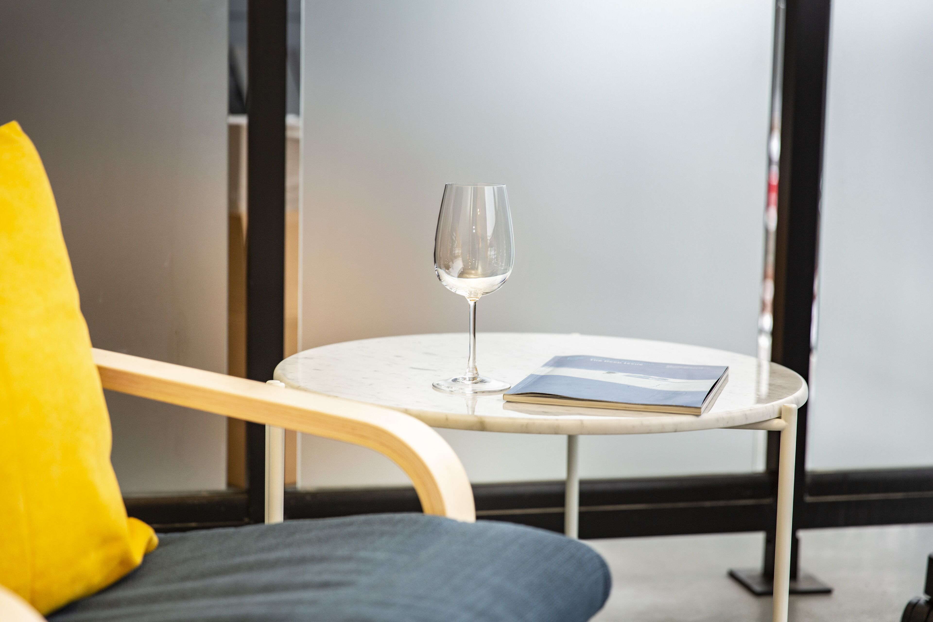 Une table ronde avec un verre de vin et un livre, un coussin jaune et une chaise avec un coussin foncé.