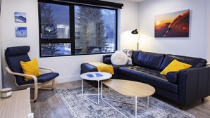 A modern living room with a dark blue leather sofa, a wooden coffee table, a blue armchair, and a large window with a view of trees and buildings. - Separate Reality By Revelstoke Vacations (Revelstoke)