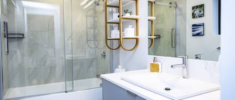 Un baño moderno con ducha de vidrio, un lavamanos blanco y un dispensador de jabón dorado, y un inodoro.
