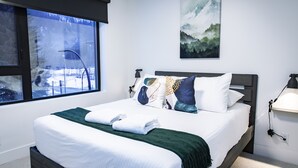 A neatly made bed with a white and green blanket, a mountain landscape painting on the wall, and a window with a view of a snowy outdoor area. - Separate Reality By Revelstoke Vacations (Revelstoke)