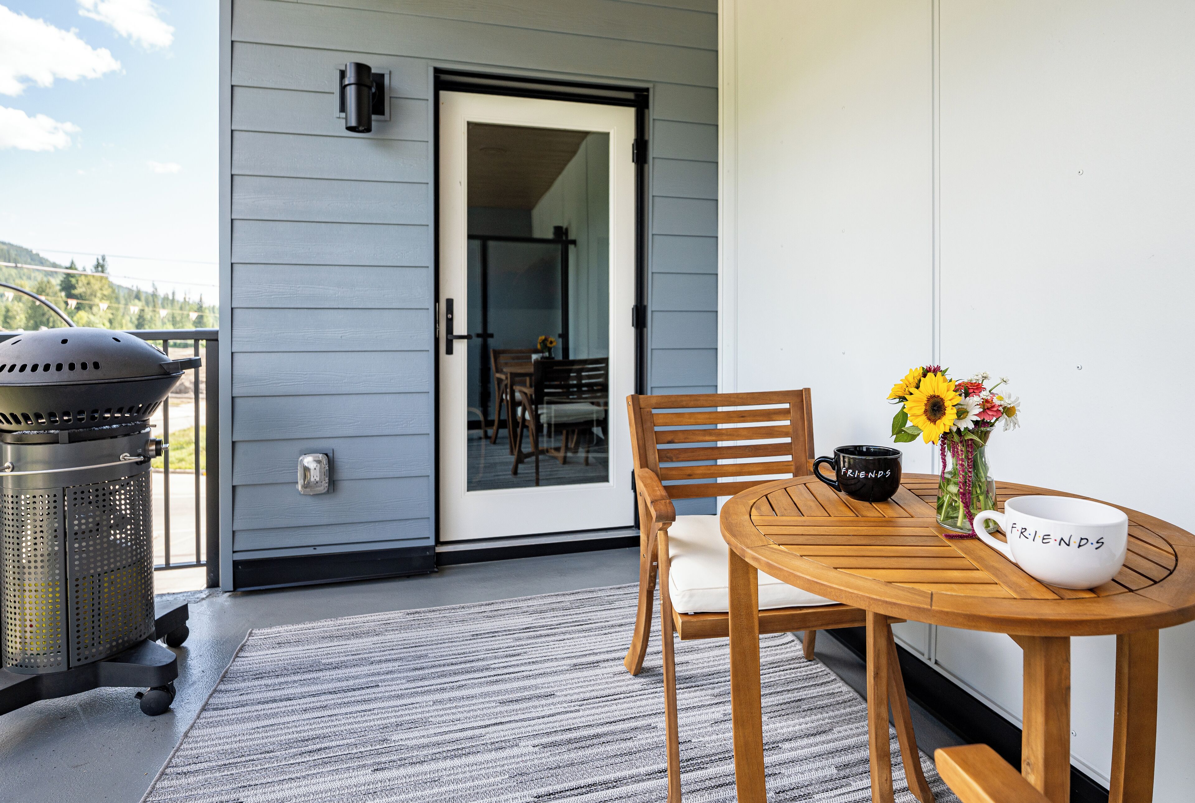 A balcony with a grill, a wooden table with chairs, a vase with flowers, and a door leading inside.