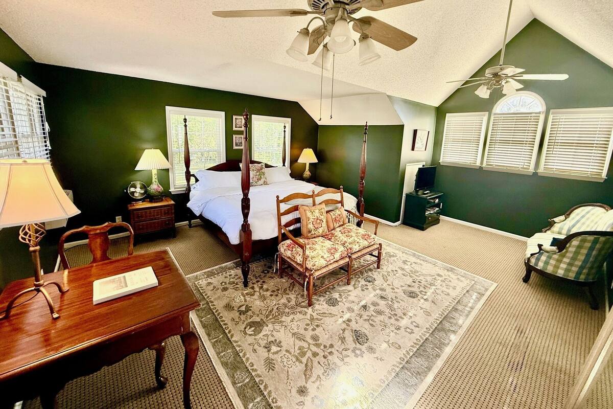 A bedroom with a four-poster bed, a desk, a chair, a ceiling fan, and a large window with blinds.