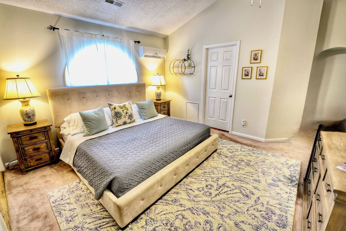 A bedroom with a large bed, two bedside tables with lamps, a window with curtains, a door, and a wall-mounted light fixture.