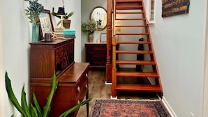A narrow hallway with a wooden staircase, a red patterned rug, and a potted plant.
