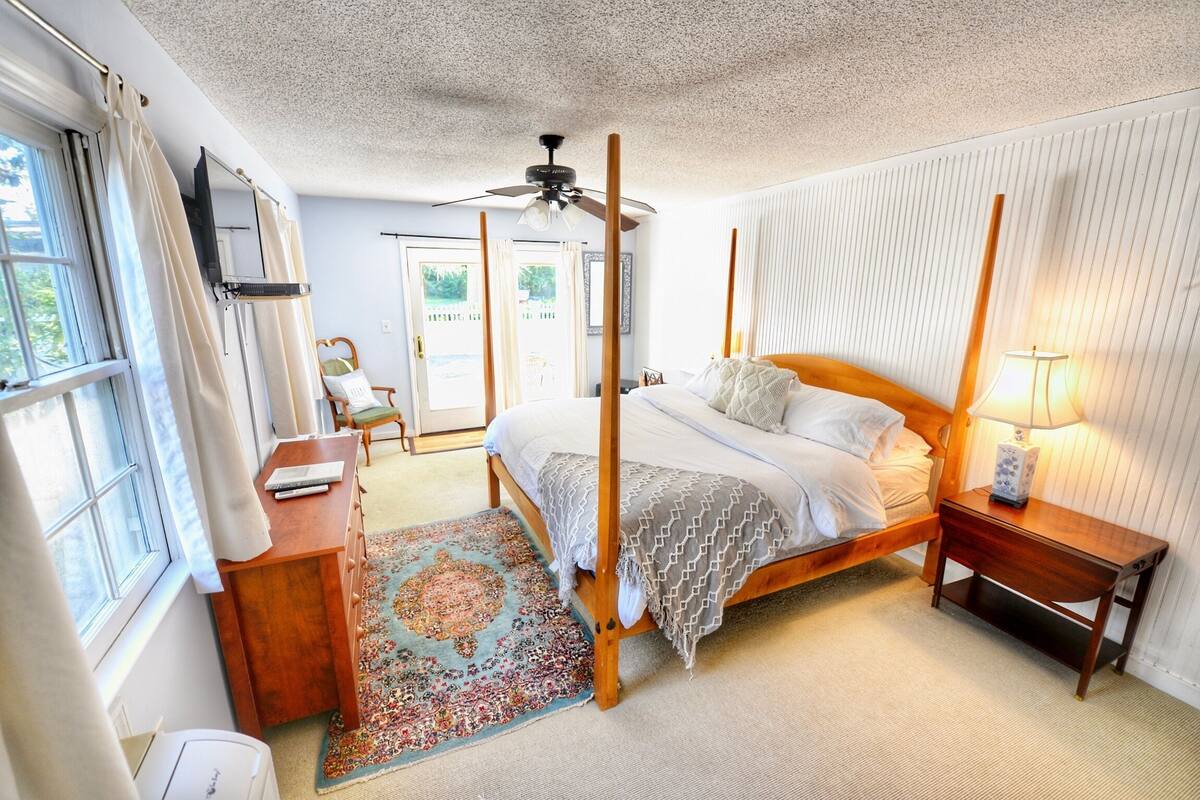 A bedroom with a four-poster bed, a wooden desk, a chair, a standing lamp, and a ceiling fan.