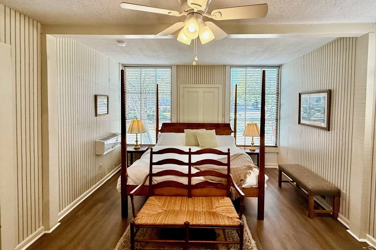 A bedroom with a four-poster bed, a bench, a ceiling fan, and two windows with blinds.