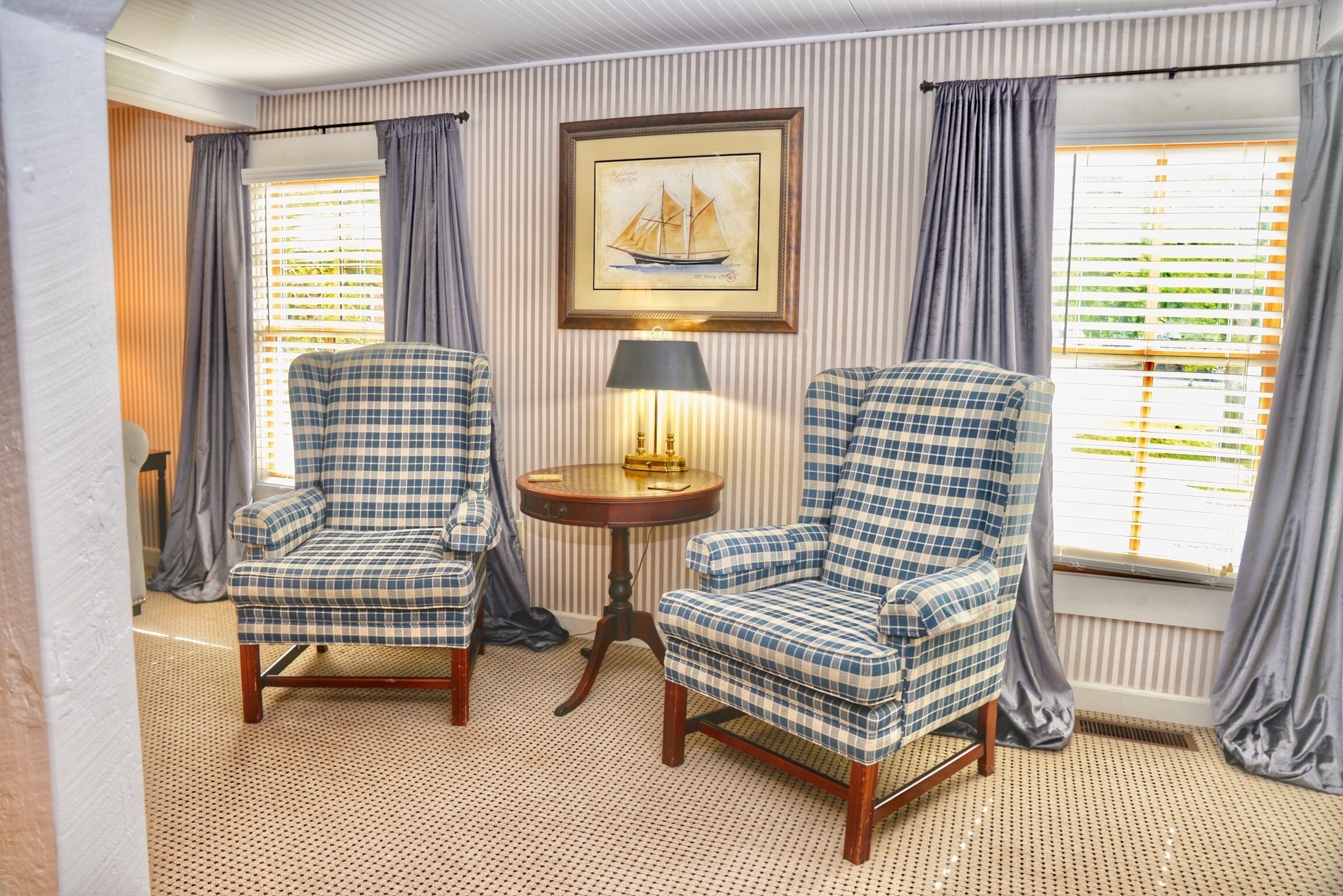 A room with two checkered armchairs, a small round table with a lamp, and a framed picture of a sailboat on the wall.
