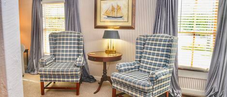 A room with two checkered armchairs, a small round table with a lamp, and a framed picture of a sailboat on the wall.