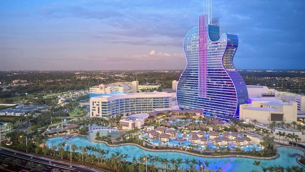 Un gran hotel con una construcción singular en forma de guitarra, múltiples piscinas y una zona de resort con palmeras.