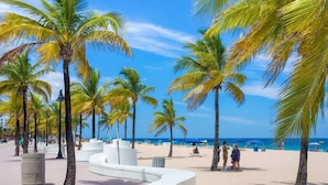 Un paseo marítimo en la playa con palmeras, un camino blanco curvado y una playa de arena con gente a lo lejos.
