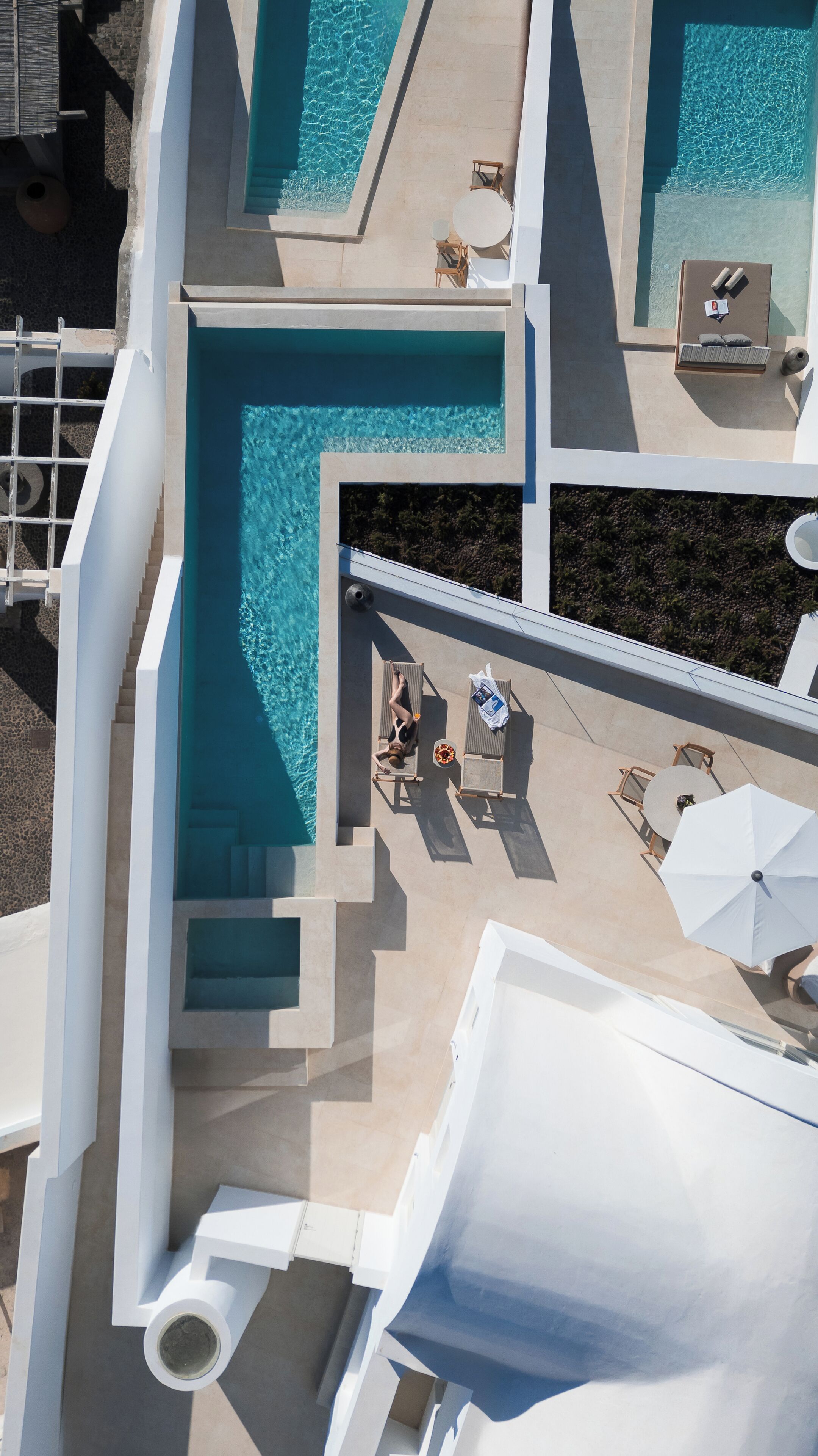 An aerial view of a modern hotel with multiple swimming pools, sun loungers, and a covered outdoor seating area.