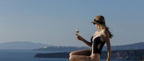 A person in a swimsuit sitting by a pool with a glass of drink, overlooking a body of water and distant mountains.