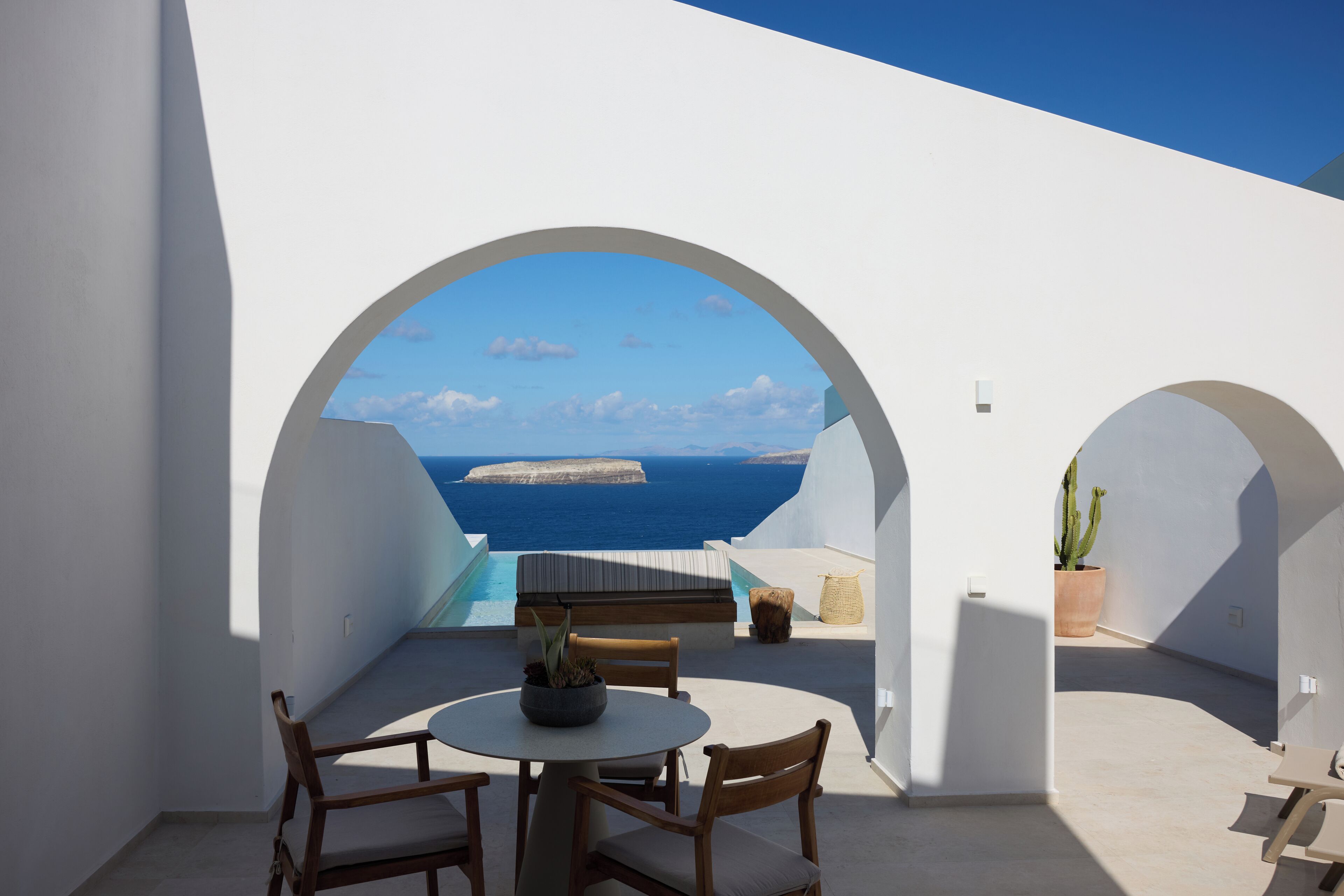 A white-walled room with a round table and chairs, a potted plant, and a view of the sea and islands.