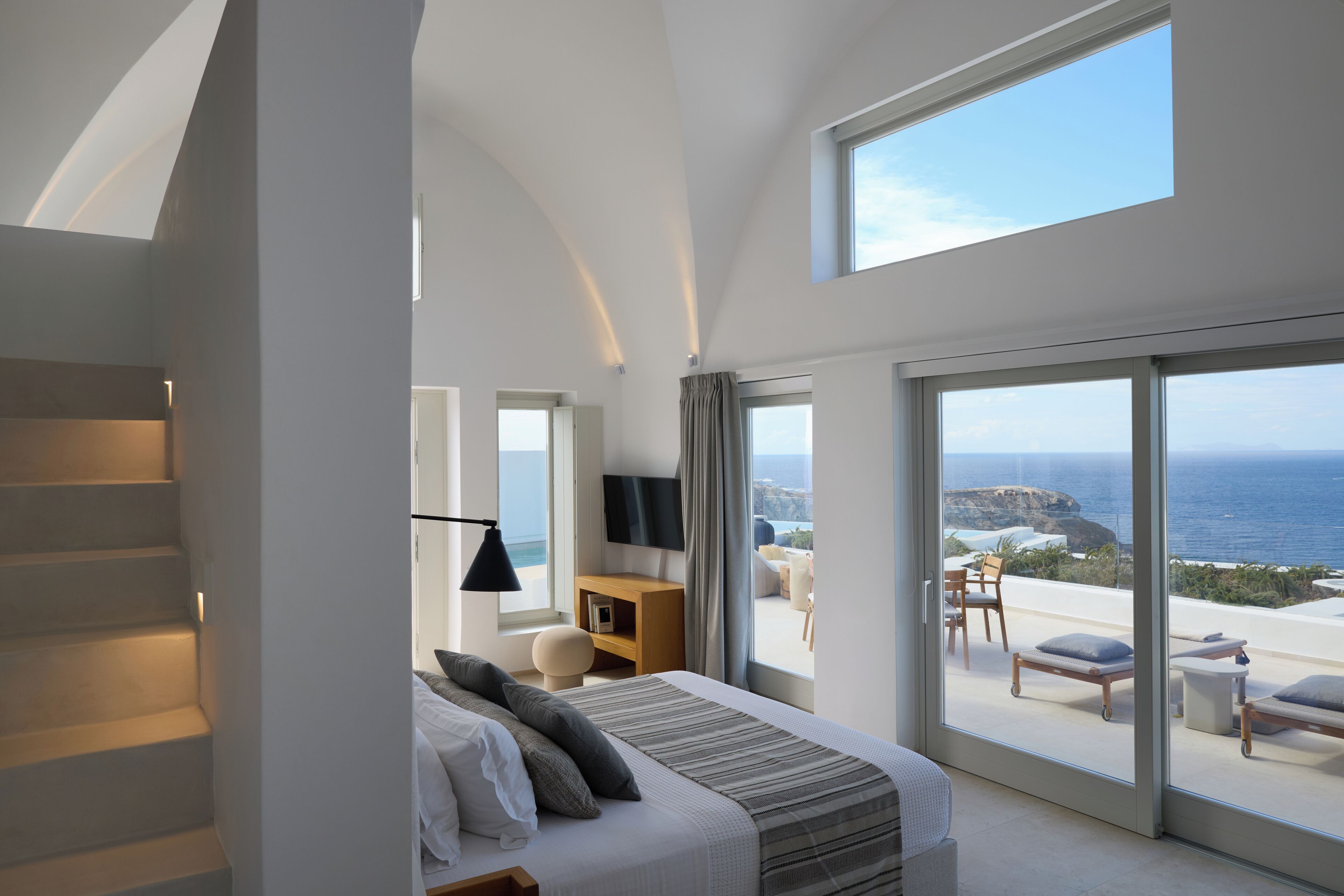 A modern bedroom with a large bed, a TV, and a balcony with a view of the sea.