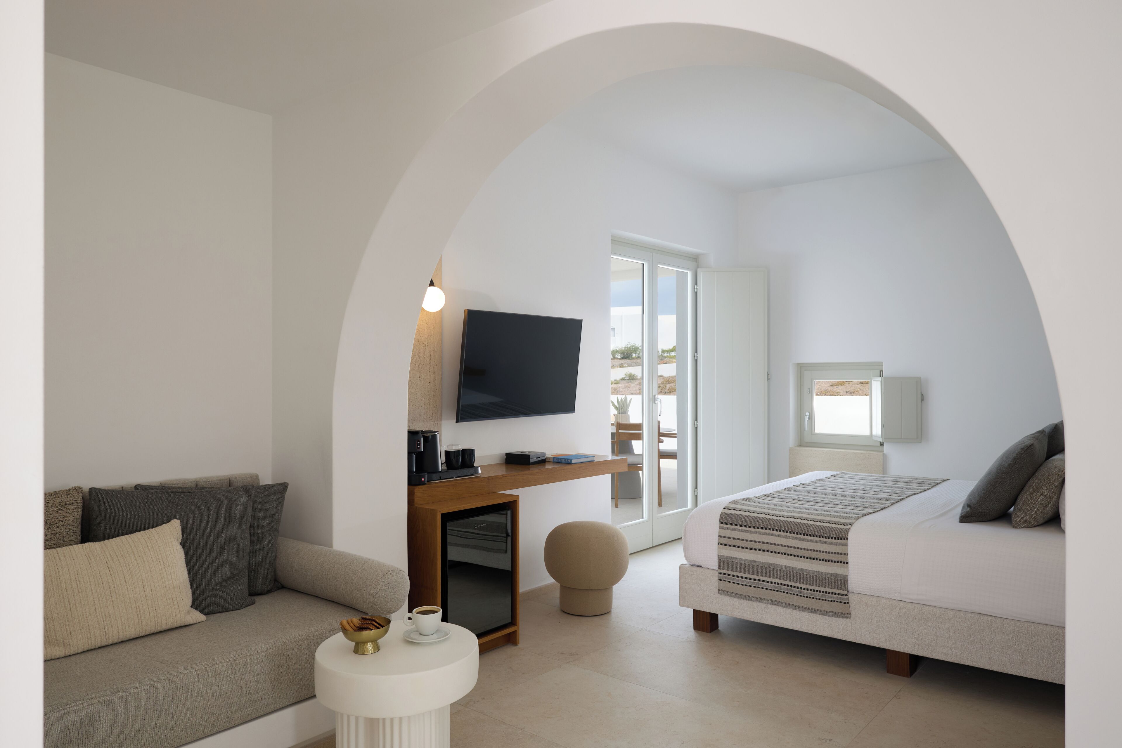 A modern, open-plan living space with a sofa, a small round table, a flat-screen TV mounted on a wooden console, and a bed with a striped blanket.