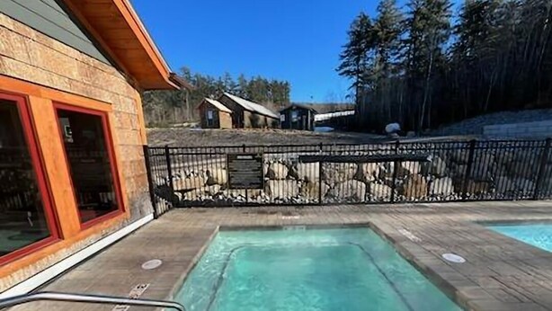 A wooden deck with a hot tub, a fenced area, and a building with a red door. - Village sunset ski in/ ski out condo on Cranmore Mountain  (Conway)