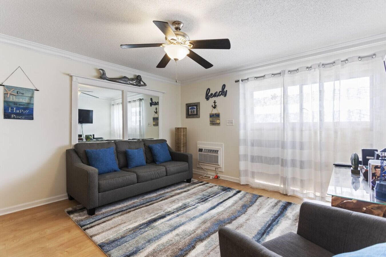 A living room with a gray sofa, blue cushions, a striped rug, a ceiling fan, a large mirror, and a window with sheer curtains.