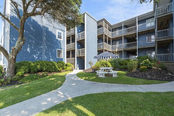 A multi-story residential building with balconies, a paved walkway, and a picnic table with an umbrella.