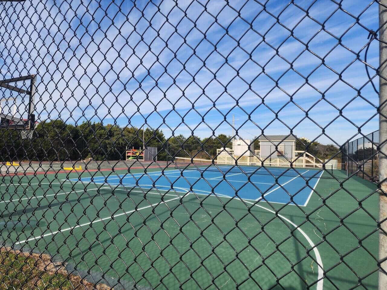 Ein Maschendrahtzaun umgibt einen Tennisplatz, im Hintergrund ist ein Gebäude zu sehen.