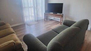 A living room with a green sofa, a brown sofa, a TV on a wooden stand, and a sliding glass door with blinds. - Shore To Please, 1st Floor Oceanfront Condo (North Topsail Beach)