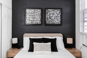 A bedroom with a dark wall, two abstract paintings, a bed with white bedding, black and white pillows, and bedside lamps.