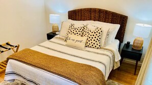 Un lit bien fait, recouvert d’une couvre-lit à rayures, d’un plaid marron et de coussins à pois. Il y a deux tables de chevet avec des lampes, et sur l’une d’elles, on aperçoit un petit panier.