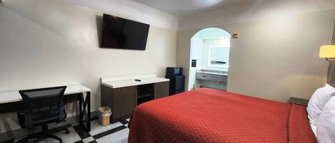 A hotel room with a bed, a desk with a chair, a television, a microwave, and a bathroom visible through an archway.