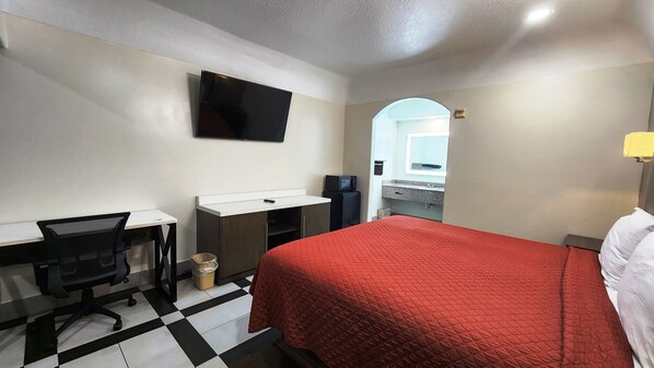 A hotel room with a bed, a desk with a chair, a television, a microwave, and a bathroom visible through an archway.
