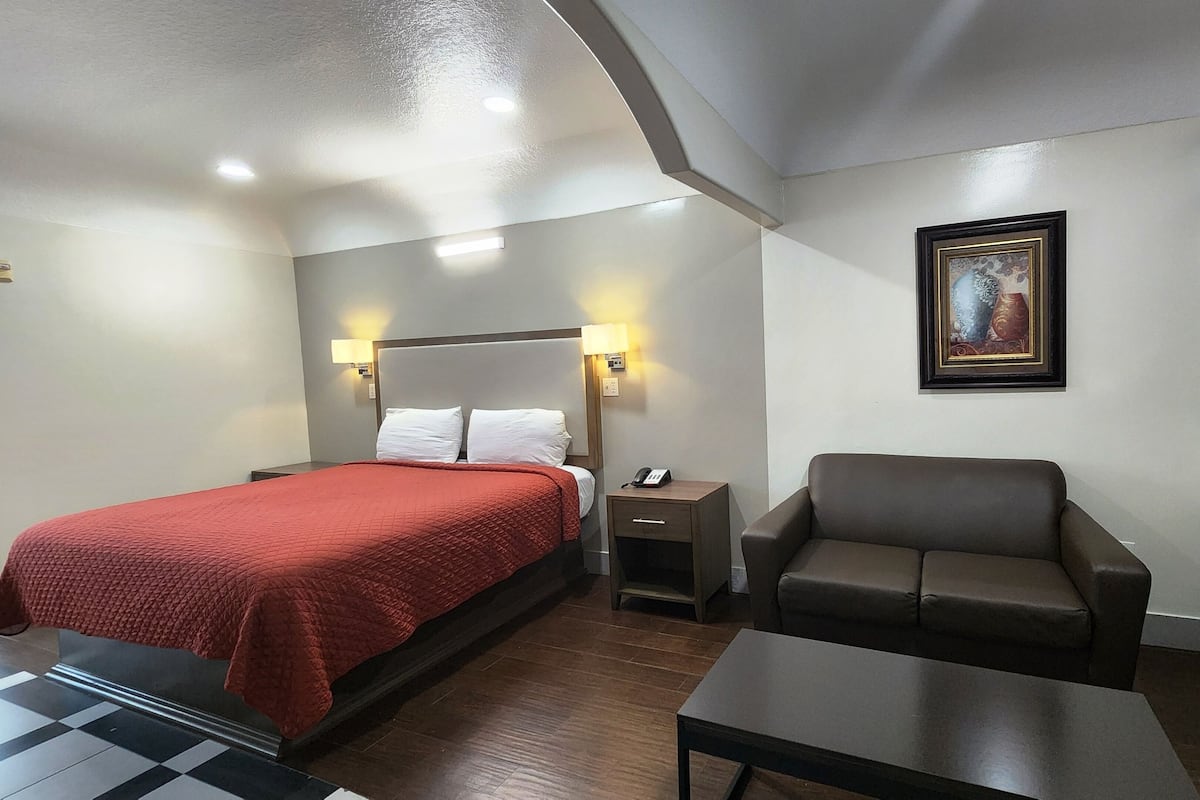 A hotel room with a bed, a chair, a small table, a nightstand, a lamp, a painting, and a window with curtains.