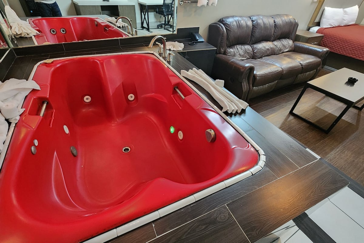 A room with a red jacuzzi tub, a leather sofa, a small table, and a bed with a red blanket.