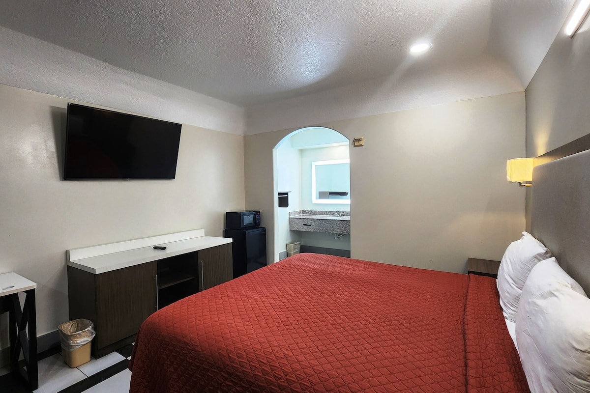 A hotel room with a bed, a desk with a computer, a TV mounted on the wall, and a small kitchenette area.