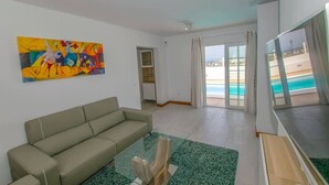 Een woonkamer met een bank, een glazen salontafel, een schilderij en uitzicht op een zwembad en strand door grote ramen.